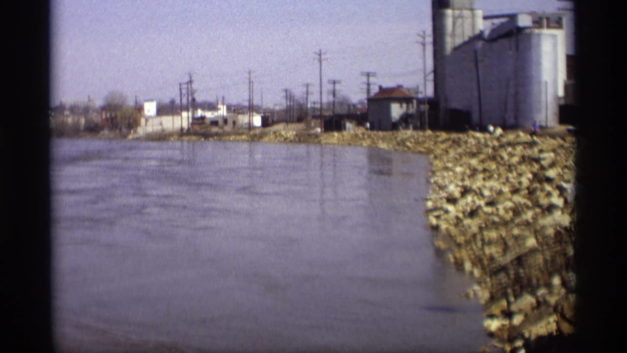 River water beside a rocky embankment with industrial tanks and utility poles in the distance.