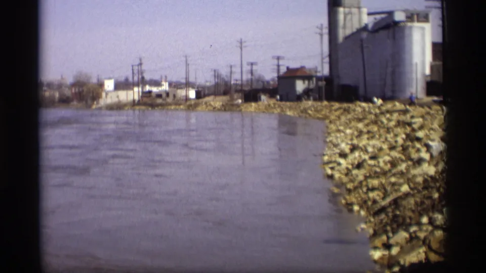 Savage, Minnesota riverbank in 1969, with moving water, riprap, industrial storage tanks, low buildings, and utility poles.