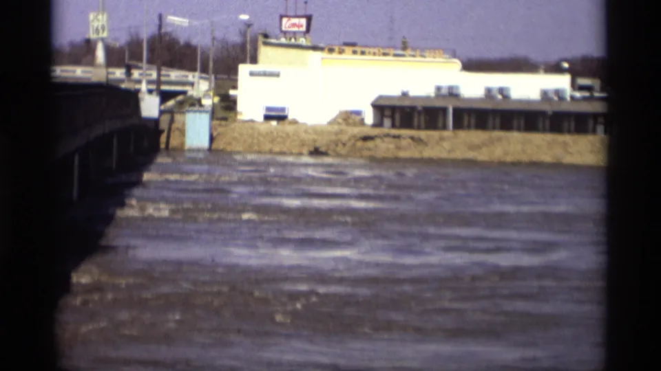 1969 8mm frame from Savage, Minnesota, looking past a bridge edge toward river water, roadside signs, and a low commercial building.