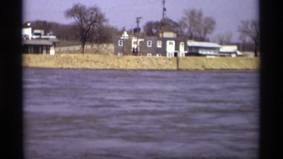Savage, Minnesota riverfront in 1969, showing moving water, bare trees, small riverside buildings, and utility poles along the bank.