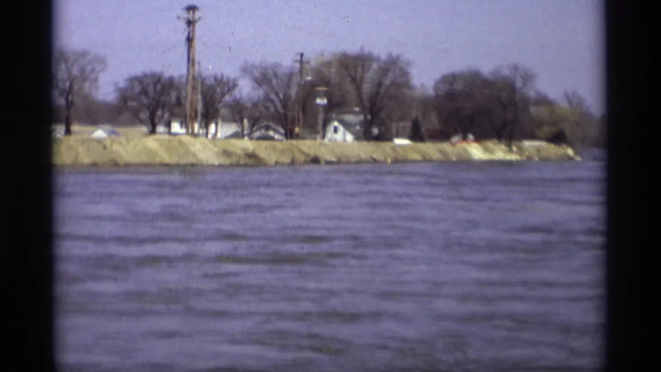 Savage, Minnesota riverbank seen from across the water, with leafless trees and small buildings in soft 8mm color.