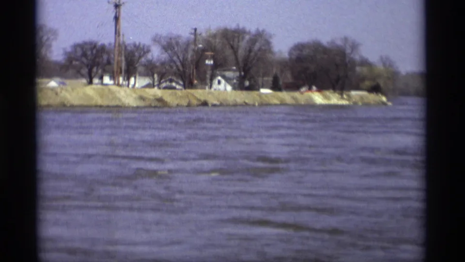 1969 home-movie frame of the Savage riverbank, with water below a grassy edge, trees, poles, and a small white building.