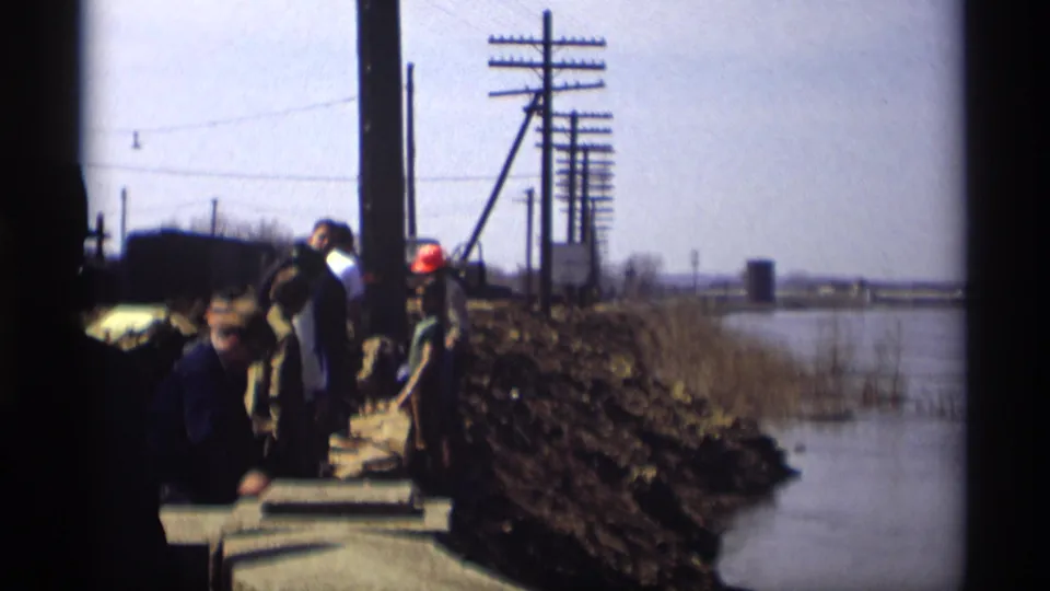 Workers in 1969 Savage, Minnesota stand along a narrow embankment beside water, with hard hats, poles, and work materials visible.