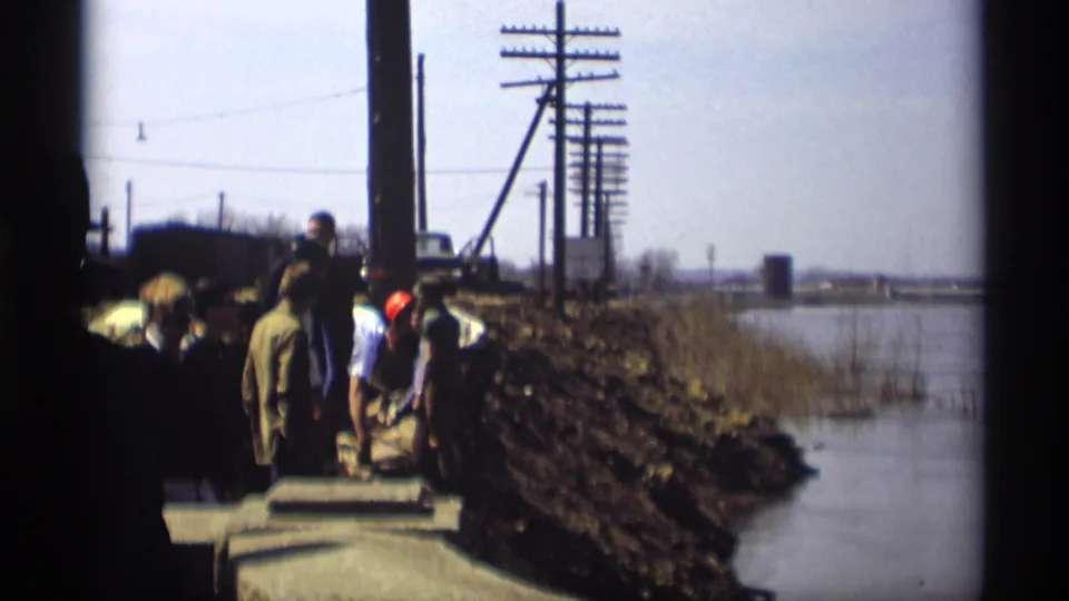 The home-movie frame shows the construction crew beside a waterside railroad embankment in Savage, Minnesota, with materials and utility poles behind them.