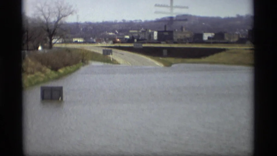 Wide 1969 Savage, Minnesota flood view with water over a road, a submerged utility box, bare trees, distant buildings, and a hillside.