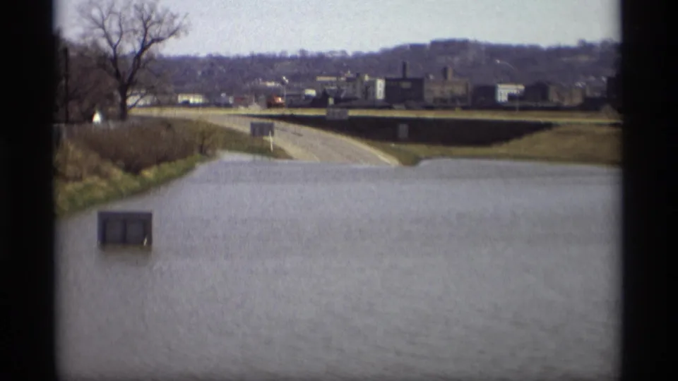 Savage, Minnesota flood scene from 1969 showing water over the roadway, a bare tree, distant low buildings, and a hillside beyond.