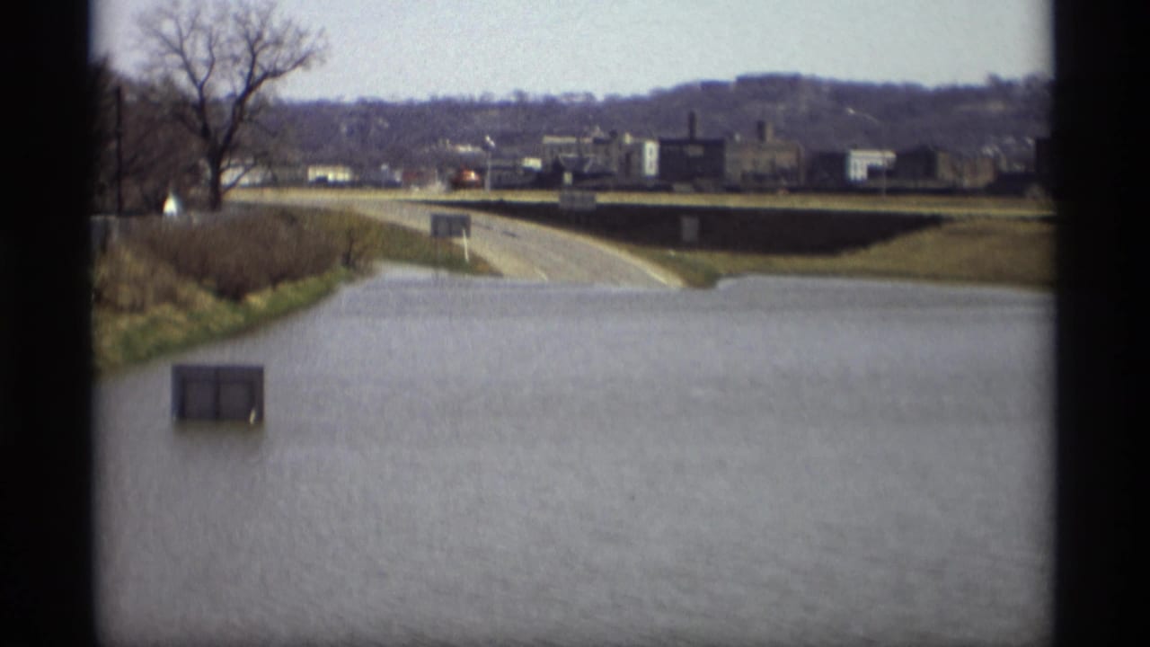 1969 Savage, Minnesota floodwater covers a roadway, with a utility box, bare trees, and distant industrial buildings visible.
