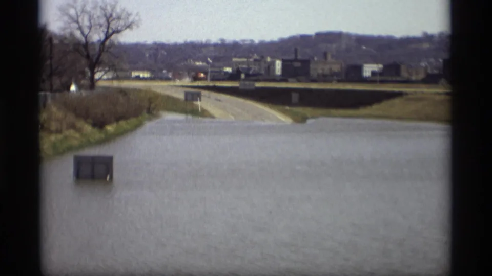 1969 Savage, Minnesota floodwater covers a roadway, with a utility box, bare trees, and distant industrial buildings visible.