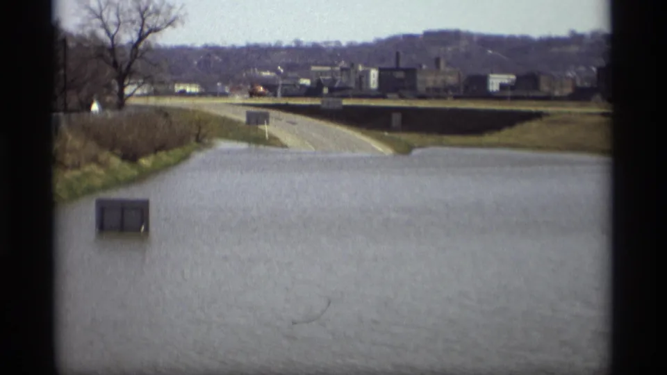 A flooded road in Savage, Minnesota appears partly submerged, with bare trees, roadside signs, and distant buildings in view.
