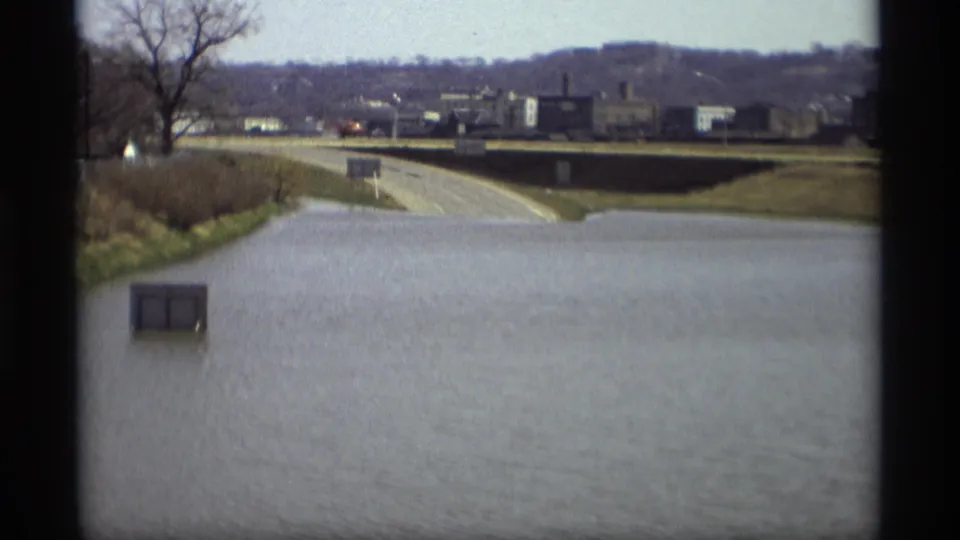 Floodwater fills the foreground in this 1969 Minnesota road scene, with a utility box, roadside signs, trees, and buildings beyond.