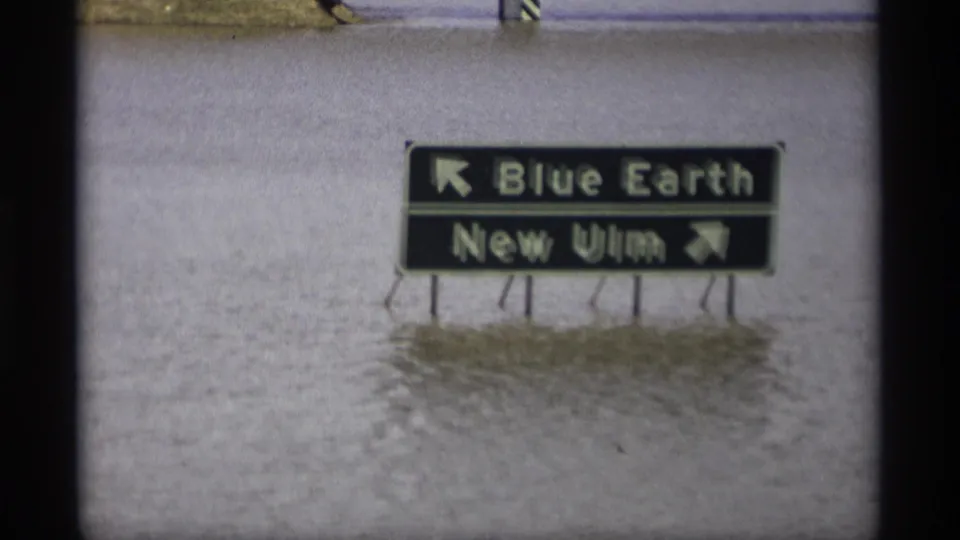 Close view of a partially submerged Minnesota directional sign for Blue Earth and New Ulm standing in floodwater.