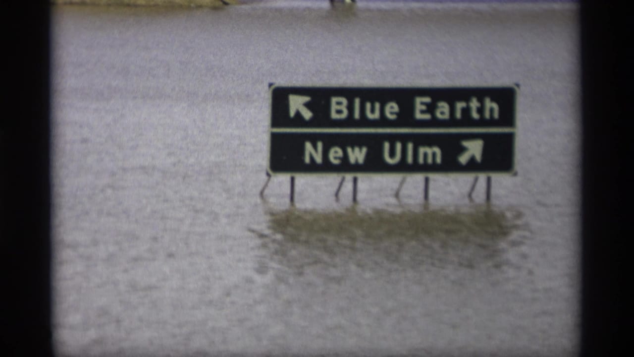 Floodwater surrounds a road sign pointing toward Blue Earth and New Ulm.