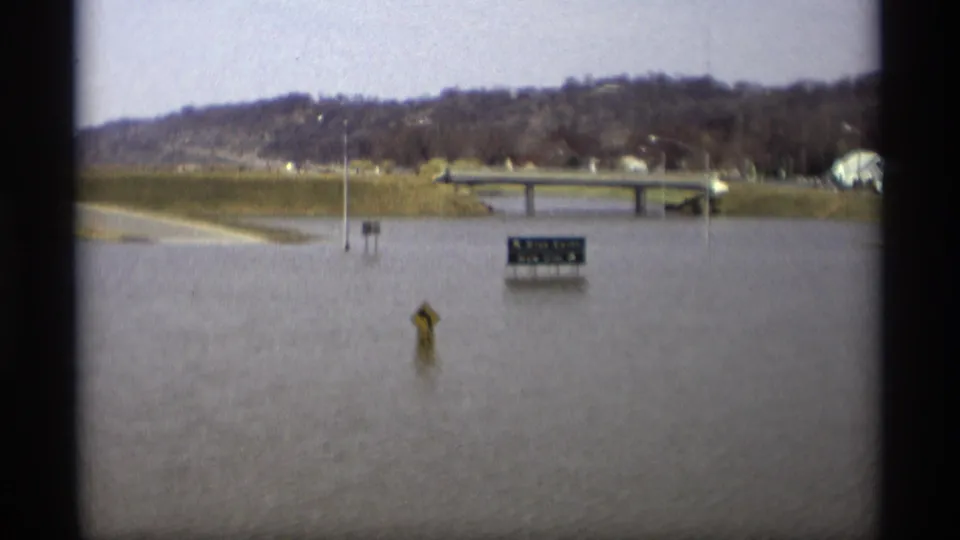 1969 Savage, Minnesota 8mm frame with floodwater across a roadway, a small bridge, and a partly readable road sign.