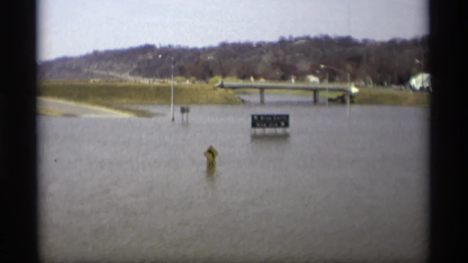 1969 Savage, Minnesota frame showing a flooded road approach, distant bridge, and directional sign in muted 8mm color.