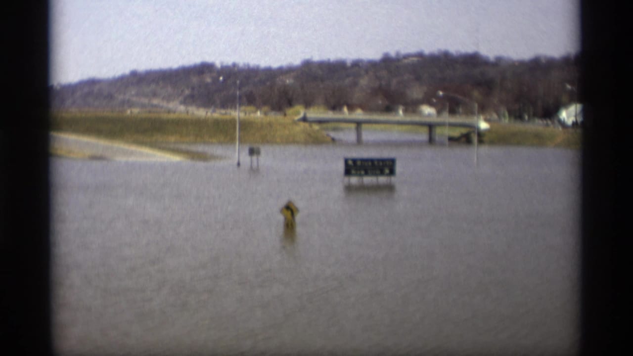 Flooded roadside area with submerged signs and a distant bridge near low hills.