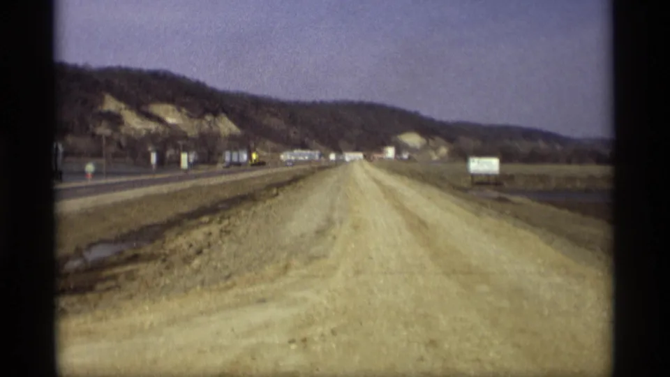 1969 Savage, Minnesota home-movie view of a dirt road beside a highway, with signboards, low wooded hills, and open fields.