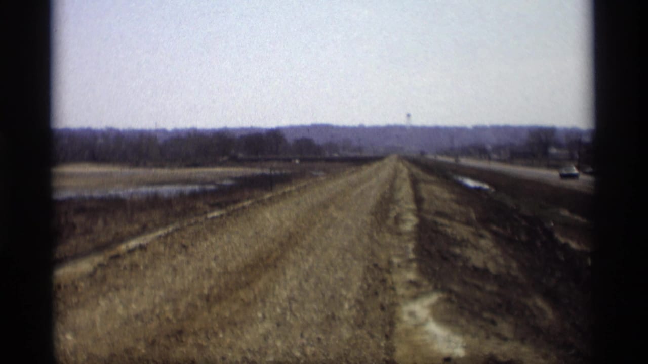 Dirt roadbed running beside a paved highway through open rural fields.
