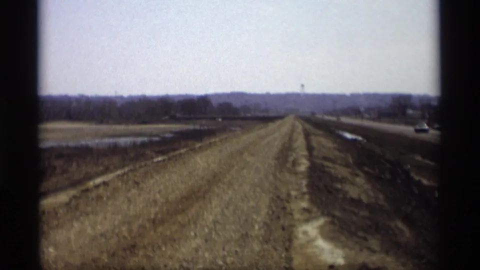 1969 Savage, Minnesota 8mm frame showing a new dirt roadbed beside a paved highway and muddy fields under a pale sky.