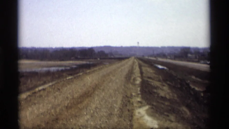 The roadbed continues through rural Savage, Minnesota, with a paved highway at right and a distant water tower near the horizon.