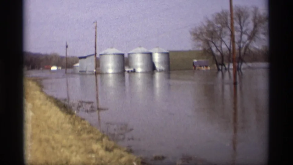 1969 Savage, Minnesota 8mm frame showing floodwater around grain silos, utility poles, bare trees, and roadside grass.
