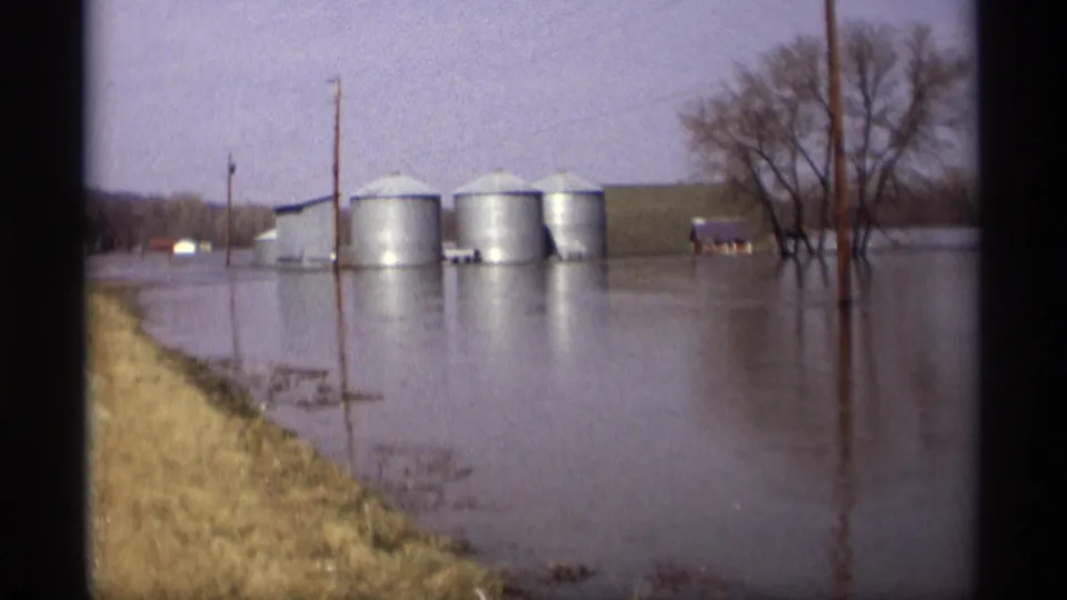 Flooded farm landscape in Savage, Minnesota, with three metal grain silos, utility poles, bare trees, and water covering low ground.