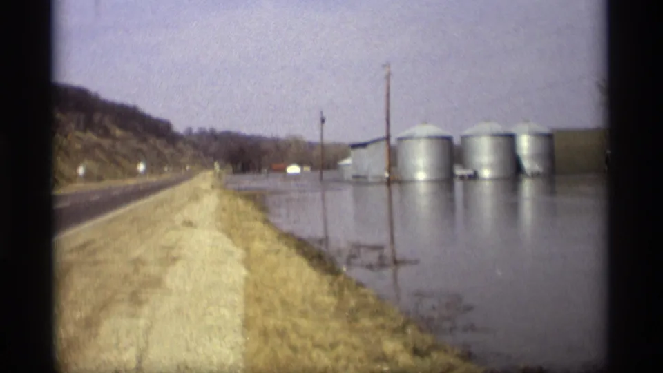 A Savage, Minnesota roadside in 1969 shows floodwater beside a highway, gravel shoulder, utility poles, farm buildings, and metal grain silos.