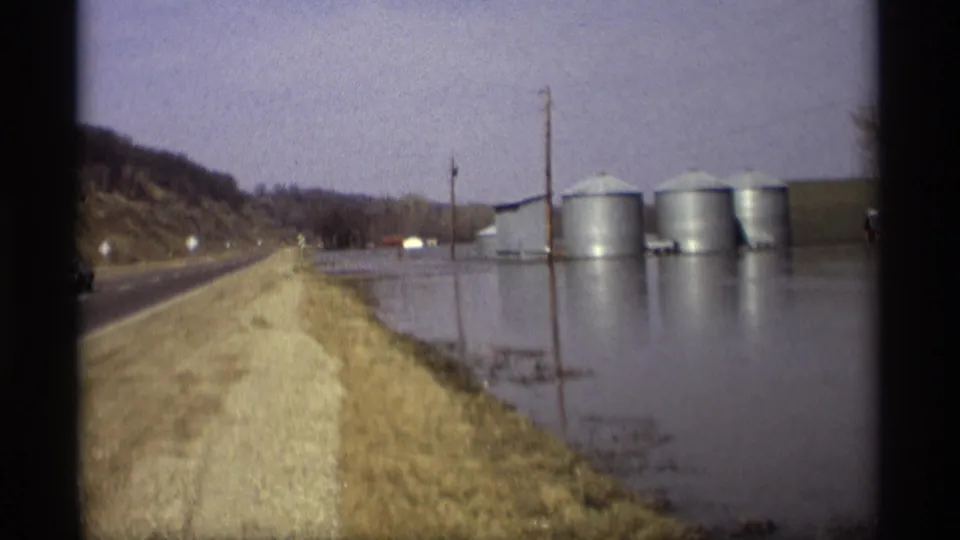 The 1969 Savage, Minnesota home-movie view follows a road past floodwater, a hillside, utility poles, and silver grain silos.