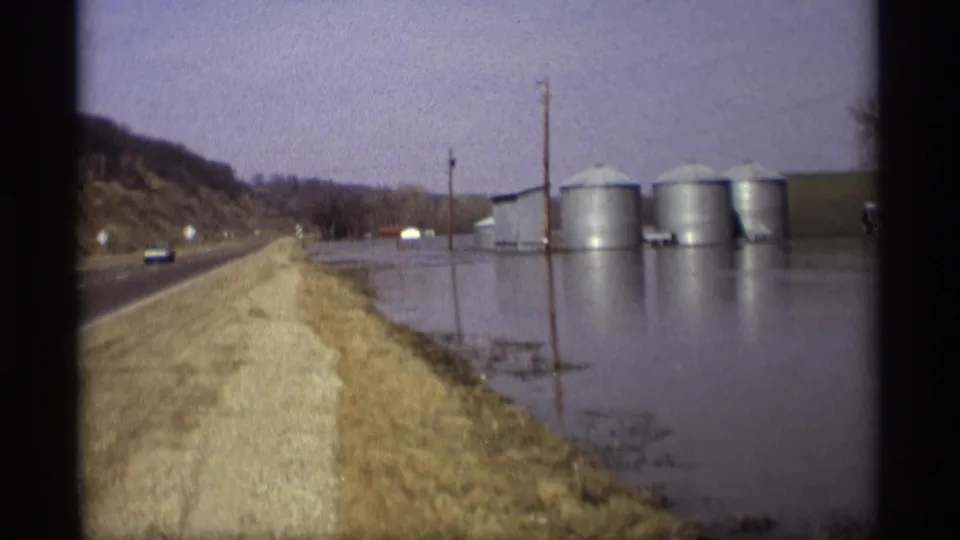 A rural Savage, Minnesota flood scene from 1969 shows a roadside, high water, farm silos, utility poles, and a small passing vehicle.