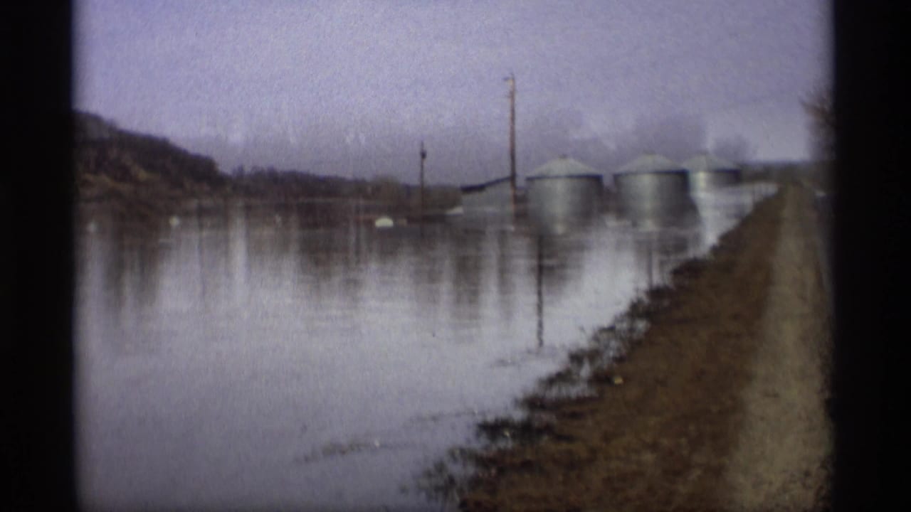 Floodwater covers farmland beside a narrow road, with grain silos and utility poles in the distance.