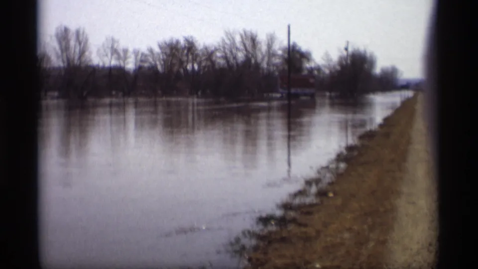1969 Savage, Minnesota frame showing floodwater along a rural road, with bare trees, a utility pole, and distant farm-adjacent structures.