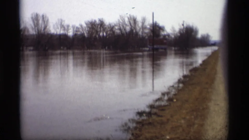 1969 Savage, Minnesota 8mm frame of floodwater along a rural road, with bare trees, utility poles, and a distant structure visible.