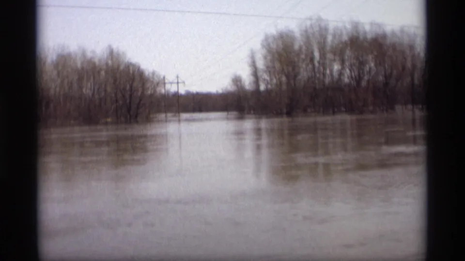 1969 Savage, Minnesota frame showing broad floodwater across low farmland or roadside land, bordered by bare trees and utility poles.