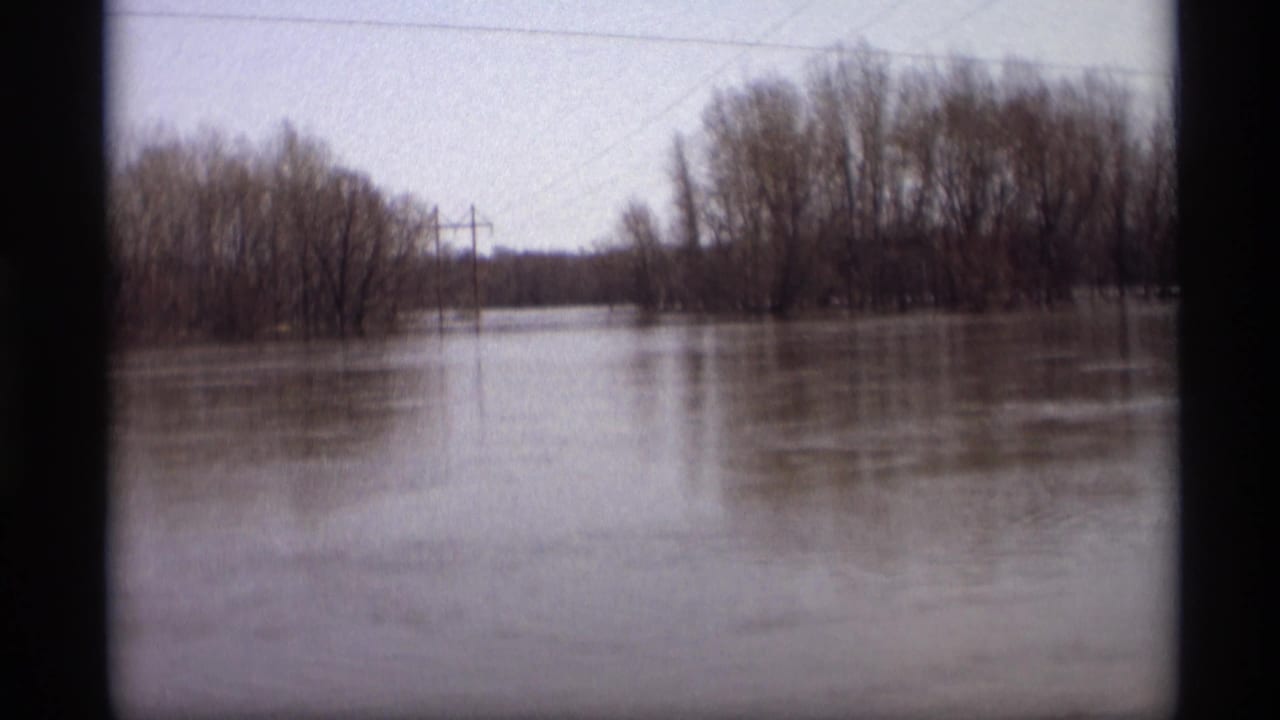 1969 8mm frame from Savage, Minnesota, showing muddy floodwater spread across a rural landscape with bare trees and utility poles.