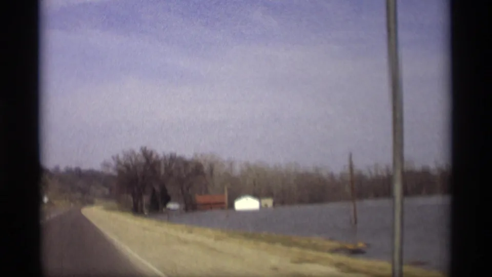 1969 Savage, Minnesota roadside view with floodwater near a road shoulder, small buildings, bare trees, and utility poles.