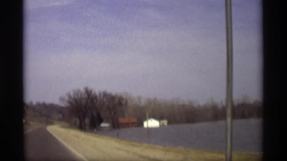 Savage, Minnesota home-movie frame showing floodwater along a road with small distant buildings and a utility pole in view.