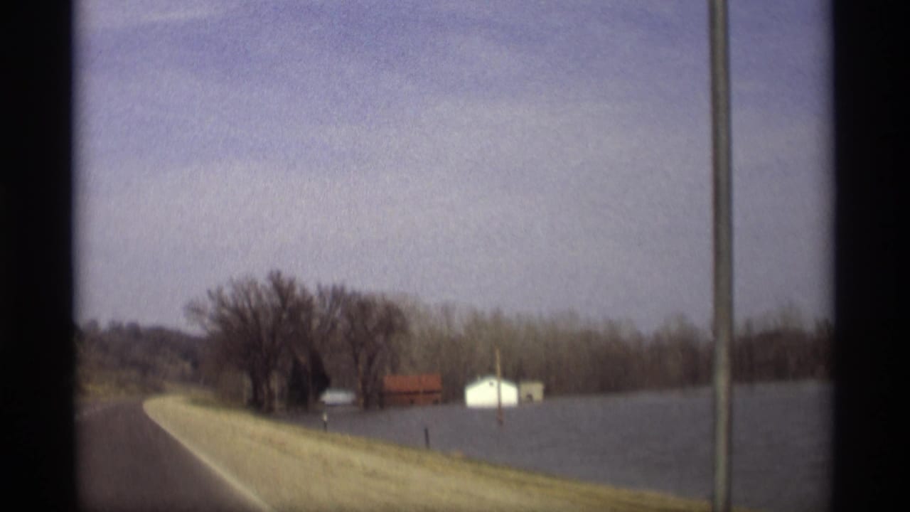 Roadway beside floodwater with bare trees and small buildings in the distance.