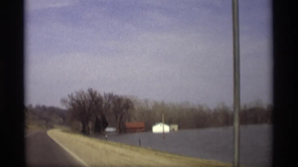 1969 Savage, Minnesota road view from a moving car, with floodwater along the shoulder and distant rural buildings.