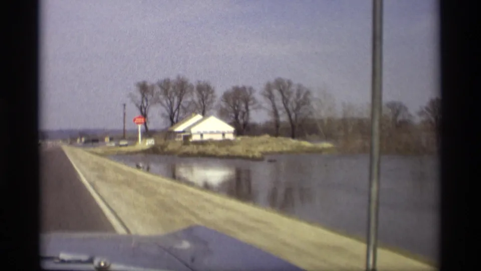 1969 Savage, Minnesota driving view approaching a red stop sign and white roadside building beside floodwater.