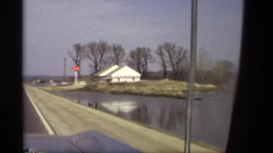Closer 1969 Savage, Minnesota road view with a stop sign, white building, bare trees, and floodwater along the roadside.