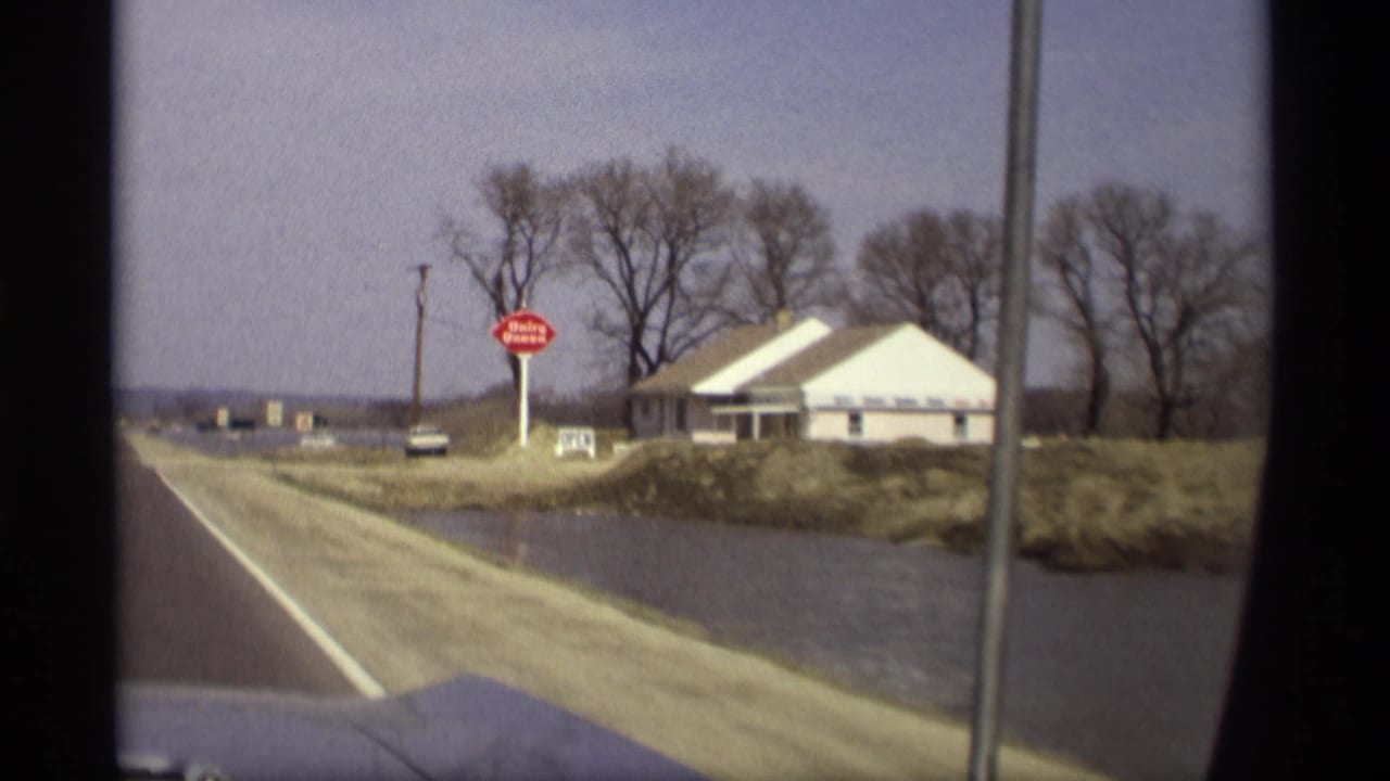 1969 Savage, Minnesota home-movie frame showing a roadside building, red sign, bare trees, and water near the road.