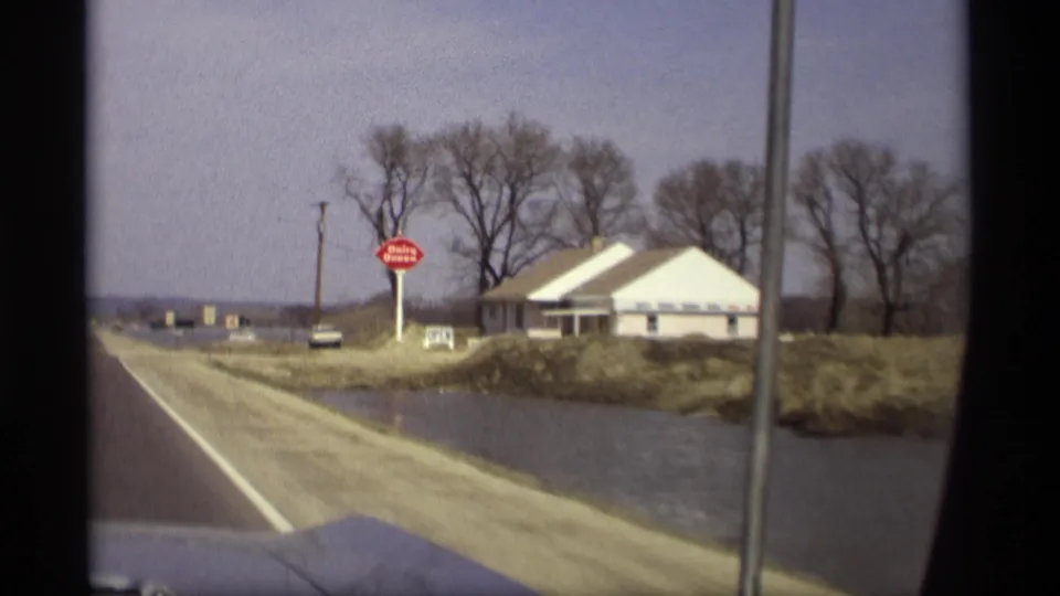 1969 Savage, Minnesota home-movie frame showing a roadside building, red sign, bare trees, and water near the road.