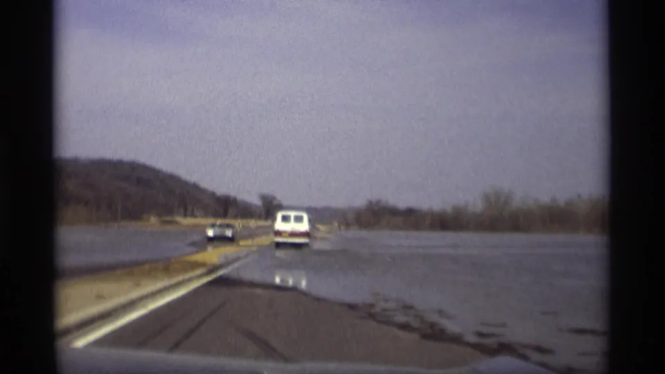 1969 Savage, Minnesota view from a moving vehicle approaching a flooded roadway with a white van and dark car ahead.