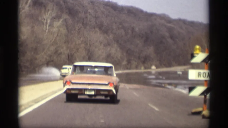 1969 Savage, Minnesota road view with a red convertible traveling near floodwater, spray, barriers, and wooded hills.