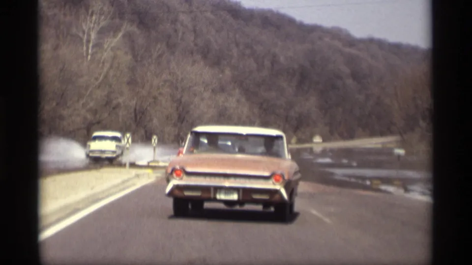 1969 Savage, Minnesota 8mm frame of a red convertible driving past roadside floodwater with trees and low hills beyond.