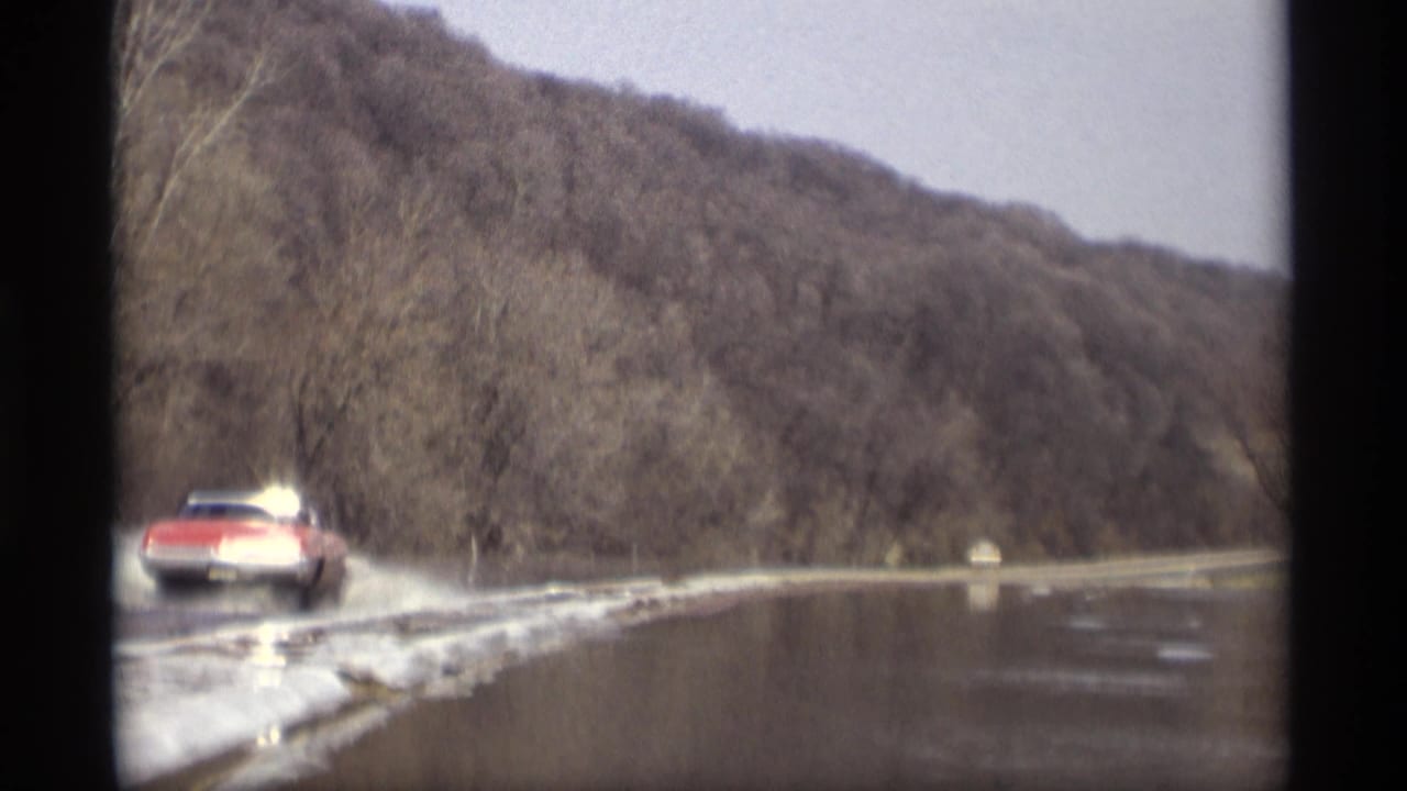 Red car drives along a flooded road beside snow and bare trees.