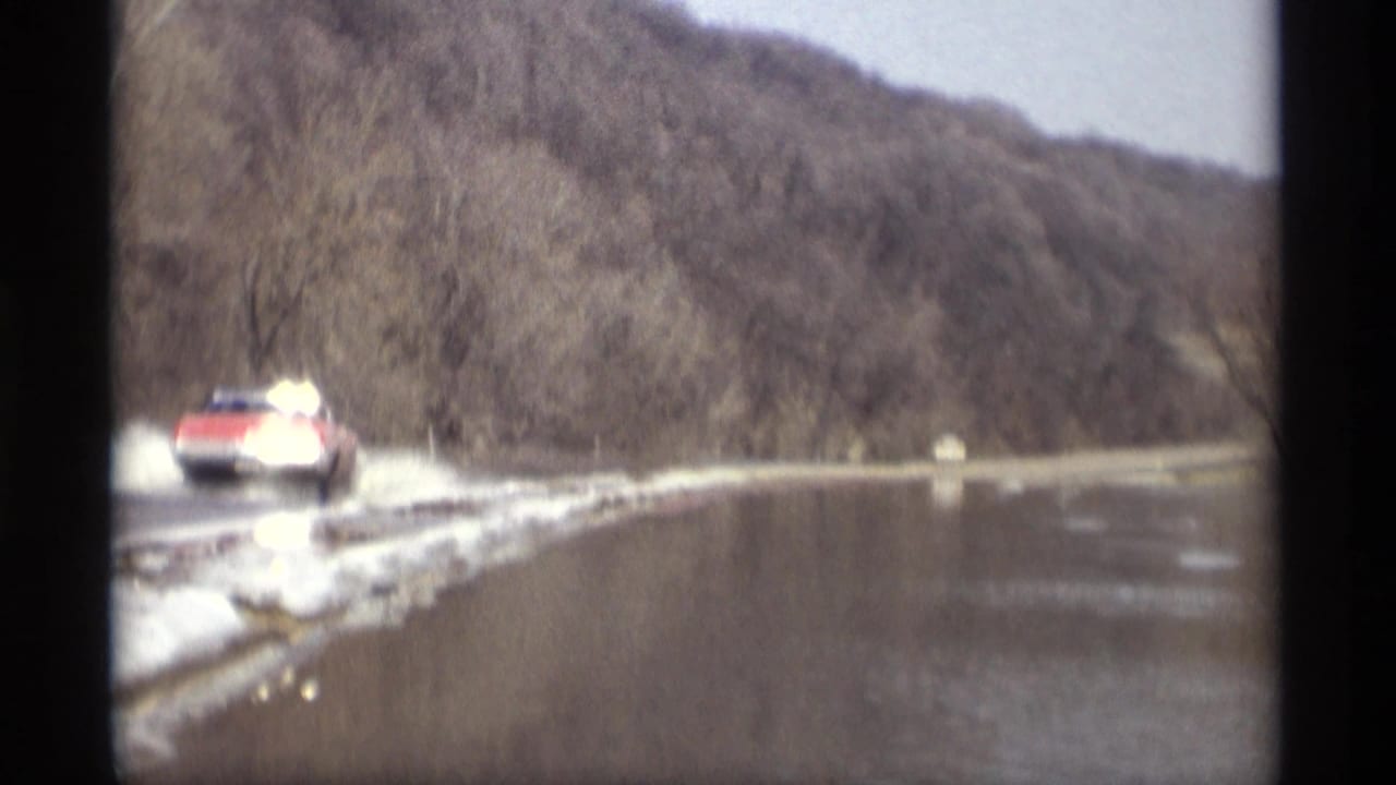 Red car splashes through water on a road with snow along the shoulder.