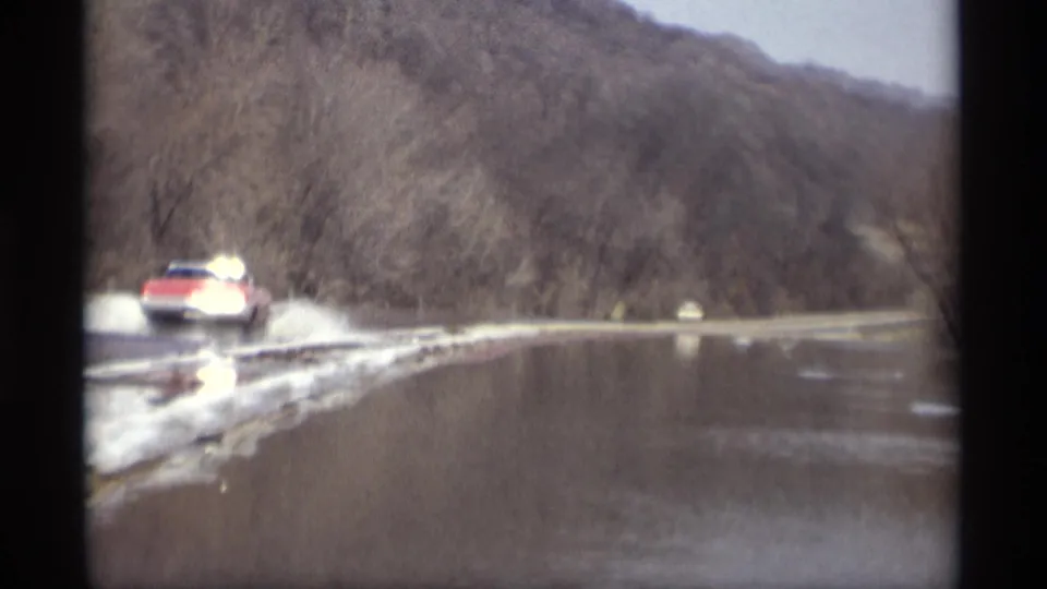 A red car continues along the flooded roadside, with reflected water, melting snow, and leafless trees visible in the frame.