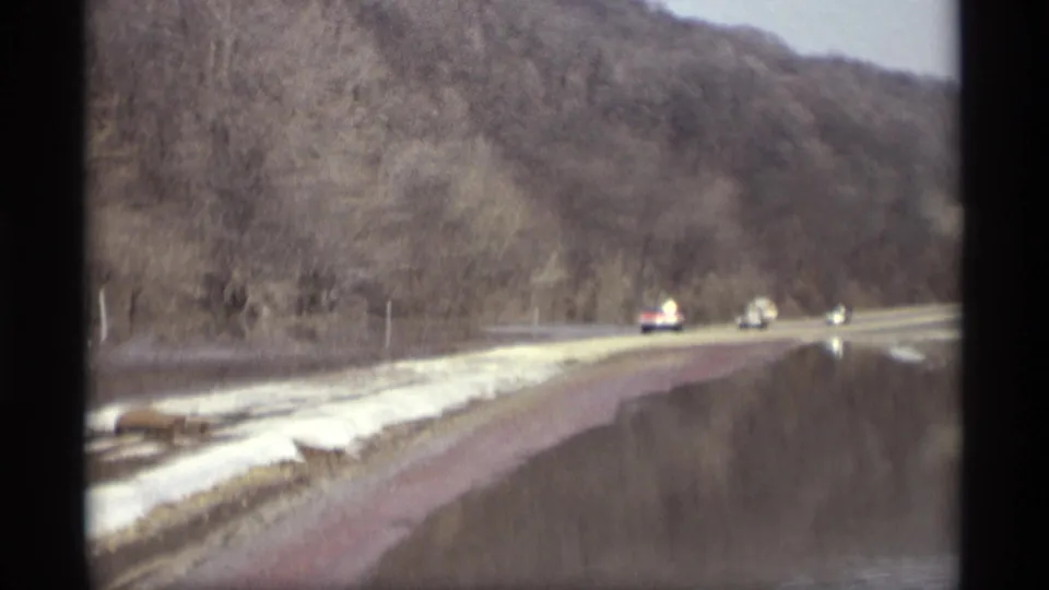 Vehicles appear farther down the wet road beside floodwater, snow patches, bare trees, and a wooded hillside in Savage, Minnesota.