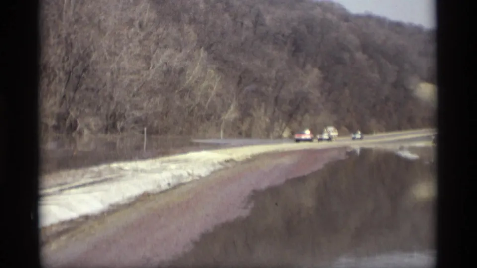 1969 Savage, Minnesota home-movie frame of cars ahead on a wet rural road bordered by water, snow, and leafless trees.