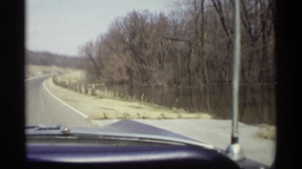 From inside a moving car, the 1969 Minnesota footage looks down a curving road bordered by water, trees, posts, and light snow.